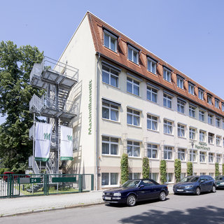 Ein mehrstöckiges Gebäude mit einem hellen, gelben Fassadenanstrich. Auf der linken Seite befindet sich ein Treppenhaus aus Metall. Im Vordergrund stehen einige parkende Autos, und es gibt Bäume in der Nähe.