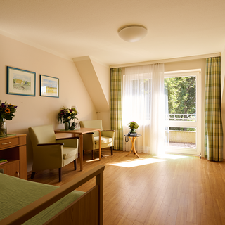 Helles und geräumiges Zimmer mit Holzfußboden. An einer Wand hängen Bilder. Links stehen ein Bett und ein Nachttisch, rechts zwei Stühle und ein Tisch. Ein offenes Fenster mit Vorhängen bietet Ausblick auf einen Balkon und die Natur draußen.