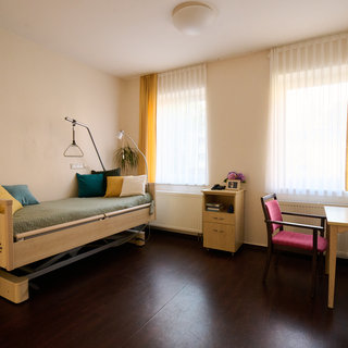 Ein heller Raum mit zwei Fenstern und langen Vorhängen. Auf der linken Seite steht ein Pflegebett mit Kissen in verschiedenen Farben. Rechts ist ein Schreibtisch mit einer pinkfarbenen Stuhl und einer Vase mit Blumen. Der Boden ist dunkel und die Wände sind neutral gefärbt.