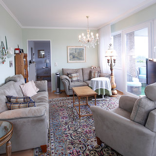 Ein gemütlicher Wohnraum mit beigen Sofas, einem Holztisch und einem Teppich. An der Wand hängt ein Bild, und ein Kronleuchter sorgt für Beleuchtung. Fenster bieten einen Blick nach draußen. Ein Stehlampe und ein Fernseher ergänzen die Einrichtung.