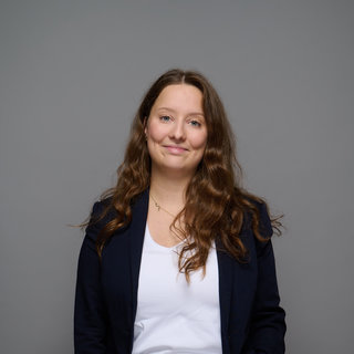 Eine junge Frau mit langen, lockigen Haaren lächelt. Sie trägt einen dunkelblauen Blazer über einem weißen T-Shirt und eine helle Hose mit floralem Muster. Der Hintergrund ist grau. Die Frau steht mit entspannter Haltung, die Hände in den Hosentaschen.