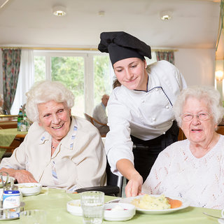 Zwei ältere Frauen sitzen an einem Tisch in einem Restaurant, beide lächeln. Eine Köchin serviert ihnen Essen. Der Tisch ist mit Geschirr und Speisen gedeckt, und im Hintergrund sind weitere Personen und eine helle, einladende Atmosphäre zu sehen.
