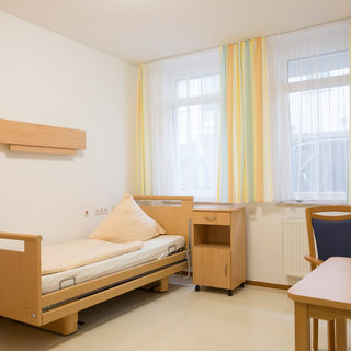 Ein einfaches Zimmer mit einem Pflegebett, das auf der linken Seite steht. Neben dem Bett befindet sich ein Nachttisch. Auf der gegenüberliegenden Seite steht ein Tisch mit einem Stuhl. Große Fenster mit hellen Vorhängen lassen viel Tageslicht herein.