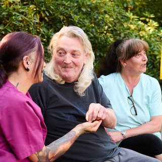 Drei Personen sitzen auf einer Bank in einem Garten. Eine Frau mit bunten Tattoos spricht lächelnd mit einem Mann mit langen Haaren, während eine andere Frau in heller Kleidung interessiert zuhört. Die Szene strahlt Freundlichkeit und Geselligkeit aus.