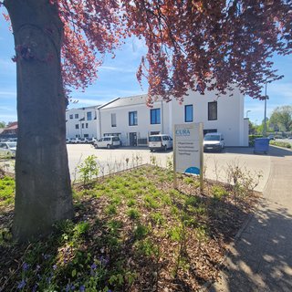 Ein helles Gebäude mit weißen Wänden und großen Fenstern ist im Hintergrund zu sehen. Im Vordergrund steht ein Schild mit der Aufschrift „CURA“ sowie Informationen zu Pflege und Wohngemeinschaften. Bunte Blumen und ein Baum mit roten Blättern umgeben die Szene.