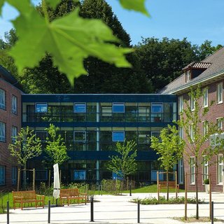 Ein modernes Gebäude mit Glasfassade befindet sich in der Mitte einer grünen Anlage. Rund um das Gebäude sind Bäume und Sträucher zu sehen. Sitzbänke laden zum Verweilen ein, und die Umgebung wirkt freundlich und einladend.