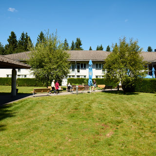 Eine grüne Wiese mit Bänken und mehreren Personen, die im Freien sitzen. Im Hintergrund sind Bäume und Gebäude sichtbar, die eine entspannte, sonnige Umgebung schaffen. Blaue Sonnenschirme stehen aufgestellt, was auf einen gemütlichen Aufenthalt hinweist.