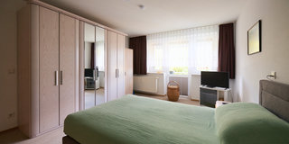 Ein Schlafzimmer mit einem großen Bett in der Mitte, bedeckt mit einem grünen Bettlaken. An der Wand steht ein großer Kleiderschrank mit Spiegeln. Neben dem Bett befindet sich ein Nachttisch. Ein Fernseher und ein Wäschekorb sind ebenfalls im Raum sichtbar. Das Licht fällt durch die Fenster.