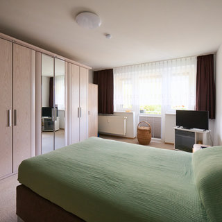 Ein Schlafzimmer mit einem großen Bett in der Mitte, bedeckt mit einem grünen Bettlaken. An der Wand steht ein großer Kleiderschrank mit Spiegeln. Neben dem Bett befindet sich ein Nachttisch. Ein Fernseher und ein Wäschekorb sind ebenfalls im Raum sichtbar. Das Licht fällt durch die Fenster.