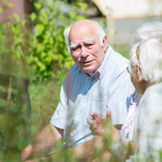 Ein älterer Mann sitzt im Freien und spricht mit einer älteren Frau. Sie scheinen sich zu unterhalten und genießen die Zeit im Garten. Die Umgebung ist grün und blühend, was eine entspannte Atmosphäre schafft.