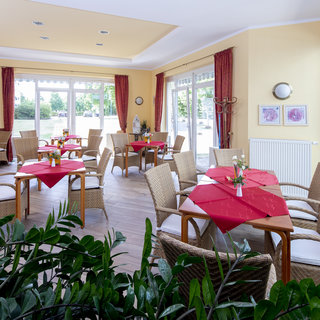 Ein helles Café mit Tischen und Stühlen aus Rattan, die mit roten Tischdecken dekoriert sind. Einige Tische sind mit Blumen geschmückt. Große Fenster lassen viel Tageslicht herein. Der Raum ist freundlich und einladend, umgeben von Pflanzen.