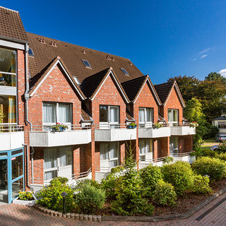 Eine Ansicht eines Gebäudes mit roter Backsteinfassade und mehreren Fenstern. Im Vordergrund sind gut gepflegte Pflanzen und ein schmaler Weg zu sehen. Der Himmel ist blau mit einigen Wolken.