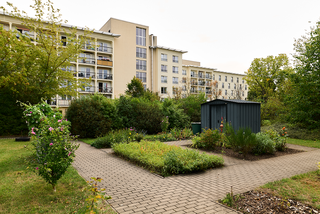 Ein Garten mit einem gepflasterten Weg, grünen Pflanzen und Blumen. Im Hintergrund steht ein Mehrfamilienhaus. Ein kleiner Geräteschuppen befindet sich in der Mitte des Gartens, umgeben von Beeten und Büschen. Der Himmel ist bewölkt.