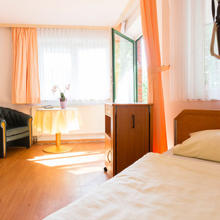 Ein heller Raum mit einem Bett im Vordergrund, einem Tisch mit einer Blume und einem Stuhl gegenüber. Große Fenster lassen Tageslicht herein, und orangefarbene Vorhänge rahmen das Fenster. Ein Holzschrank steht neben dem Tisch. Der Raum wirkt einladend und freundlich.