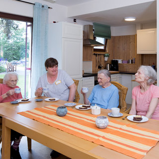 Vier ältere Frauen und eine Betreuerin sitzen an einem Tisch in einer hellen Küche. Die Frauen halten Tassen in der Hand und essen Kekse. Es herrscht eine freundliche Atmosphäre während des gemeinsamen Nachmittags.