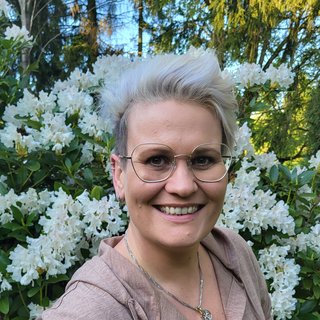 Eine Person mit kurzen, blonden Haaren und einer Brille lächelt freundlich vor einem Hintergrund aus blühenden, weißen Rhododendren. Sie trägt einen beigen Pulli mit Knöpfen und hat eine Kette mit einem Anhänger um den Hals. Der Gesichtsausdruck ist fröhlich und einladend.