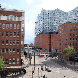 Blick auf eine moderne Innenstadt mit einer Mischung aus Backsteingebäuden und einer auffälligen, geschwungenen Glasarchitektur im Hintergrund. Auf der Straße sind Fußgänger und Fahrzeuge sichtbar, während eine Brücke über einen Gehweg führt. Der Himmel ist teilweise bewölkt.
