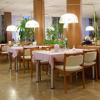 Ein Restaurantbereich mit Holztischen, die mit rosa Tischdecken und Stühlen ausgestattet sind. Es gibt mehrere runde Lampen über den Tischen, und im Hintergrund sind Pflanzen sichtbar. Einige Gäste sind an den Tischen und genießen ihre Mahlzeit.