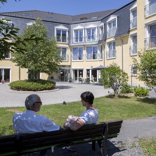 Zwei Personen sitzen auf einer Bank im Freien und schauen auf ein Wohngebäude. Es ist ein sonniger Tag, umgeben von Bäumen und Sträuchern. Die Atmosphäre wirkt entspannt und freundlich, ideal für ein Gespräch im Freien.