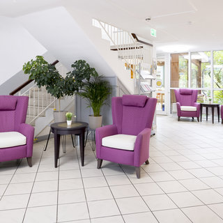 In einem hellen Raum stehen vier lila Sessel mit weißen Kissen um einen kleinen Tisch. Eine Pflanze steht in einer Ecke. Im Hintergrund sind Treppen und große Fenster, die Tageslicht hereinlassen. Der Boden ist mit hellen Fliesen ausgelegt.