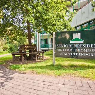 Ein Schild mit der Aufschrift "Seniorenresidenz 'Unter der Homburg'" steht vor einem Gebäude. Im Vordergrund ist ein gepflasterter Weg zu sehen, der von Bäumen umgeben ist. Bankplätze laden zur Erholung im Freien ein.
