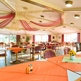 Der Raum zeigt ein helles, einladendes Speiselokal mit Tischen und Stühlen. Die Tische sind mit orangenen und grünen Tischdecken dekoriert. Über dem Raum hängen leichte Stoffe. Pflanzen und Fenster sorgen für eine angenehme Atmosphäre. Es ist ein gemütlicher Ort zum Essen und Verweilen.