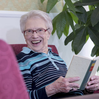 Eine ältere Frau sitzt in einem Sessel, lächelt und hält ein geöffnetes Buch in den Händen. Sie trägt eine gestreifte Bluse und eine Brille. Im Hintergrund sind Pflanzen zu sehen, die eine gemütliche Atmosphäre schaffen.