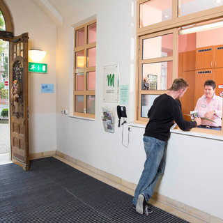 Eine Person steht an einem Empfangsfenster und spricht mit einer Mitarbeiterin. Die Empfangsdame sitzt hinter dem Fenster und lächelt, während sie Dokumente entgegennimmt. Der Raum ist hell erleuchtet und hat eine einladende Atmosphäre.