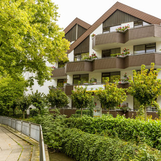 Eine moderne Wohnanlage mit drei Dachgauben, umgeben von üppigem Grün. Auf den Balkonen stehen Pflanzen. Ein gepflasterter Weg führt entlang eines kleinen Baches, der von Sträuchern gesäumt ist. Die Szene vermittelt eine ruhige und einladende Atmosphäre.