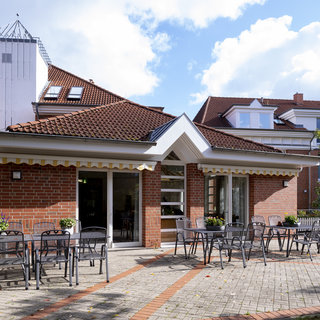 Das Bild zeigt ein modern gestaltetes Gebäude mit roten Ziegelwänden und einem Satteldach. Vor dem Gebäude befinden sich mehrere Tischgruppen mit Stühlen auf einem gepflasterten Bereich. Im Hintergrund sind weitere Gebäude und Bäume sichtbar, unter einem klaren blauen Himmel.