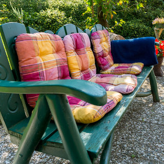 Eine grüne Holzbank im Freien ist mit bunten Kissen in Gelb, Rot und Lila ausgestattet. Diese Kissen sind zur bequemeren Nutzung der Bank angeordnet. Im Hintergrund sind Pflanzen und eine kleine Blumenbank sichtbar. Das Bild vermittelt eine einladende, entspannte Atmosphäre im Garten.