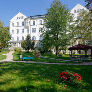 Ein grüner Garten mit Bäumen und Blumen umgibt ein großes, helles Gebäude. Im Vordergrund sind Bänke und ein Pavillon sichtbar, die zum Verweilen einladen. Der Himmel ist blau und klar, was eine freundliche Atmosphäre schafft.