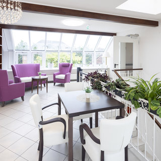 Ein heller Raum mit großen Fenstern, bestückt mit einem Tisch und Stühlen. Zwei lila Sessel stehen neben dem Tisch, umgeben von Pflanzen. Das Design vermittelt eine einladende und gemütliche Atmosphäre.