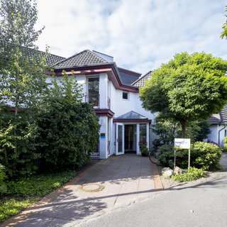 Ein modernes Haus mit einem weißen Außenanstrich und einem roten Dach. Der Eingangsbereich ist lichtdurchflutet und hat eine Glastür. Rundherum sind Bäume und Sträucher, die eine grüne Umgebung schaffen. Ein Weg führt zur Haustür.