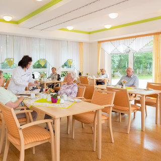 Eine helle, einladende Cafeteria mit mehreren Tischen. Eine Angestellte serviert Getränke an älteren Menschen, die entspannt am Tisch sitzen. Fenster bieten Blick auf den Garten. Blumen auf den Tischen schaffen eine freundliche Atmosphäre.