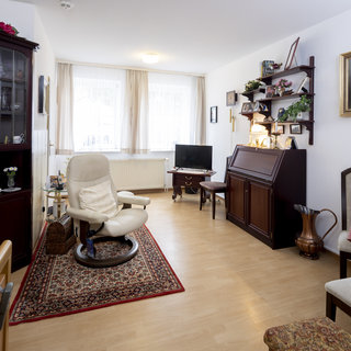 Ein gemütlicher Wohnraum mit hellem Holzfußboden. Auf der linken Seite steht ein Bücherregal und ein Stuhl mit einem runden Teppich. Rechts ist ein Klavier, ein Tisch und Stühle. Ein Fernseher ist ebenfalls sichtbar, und große Fenster lassen viel Tageslicht herein.