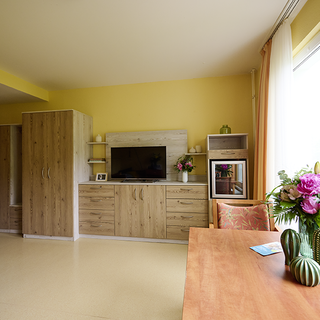 Ein heller Raum mit gelben Wänden, ausgestattet mit einem Holzschrank, einem Fernsehschrank und einem großen Fenster. Auf einem Tisch steht eine Blumenvase mit bunten Blumen, daneben ein kleiner Kaktus und ein Tischgedeck. Ein Sofa ist links im Bild sichtbar.