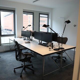 Ein modernes Büro mit mehreren Schreibtischen und Doppelmonitoren. Die Arbeitsplätze sind ausgestattet mit Schreibtischlampen und Bürostühlen. Durch große Fenster fällt Tageslicht in den Raum, der einen Blick auf ein angrenzendes Gebäude ermöglicht. Der Boden ist mit Teppich ausgelegt.