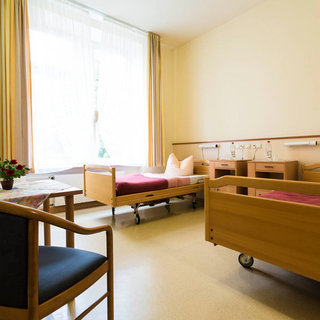 Ein heller, freundlicher Raum mit zwei Betten, einem Tisch und einem Stuhl. Die Betten sind mit Decken und Kissen ausgestattet. Am Fenster sind helle Vorhänge angebracht, und es gibt einen kleinen Tisch mit einer Blumenpflanze.