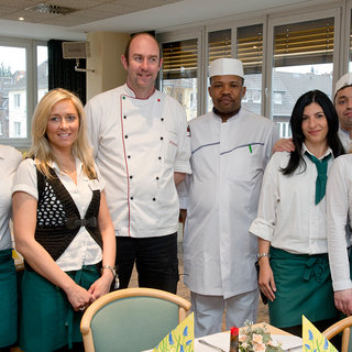Eine Gruppe von sechs Personen steht in einem gut beleuchteten Restaurant. Sie tragen einheitliche Kleidung: die Kellnerinnen haben weiße Blusen und grüne Schürzen, während der Koch eine weiße Uniform und eine Mütze trägt. Sie lächeln und scheinen stolz auf ihre Arbeit zu sein.