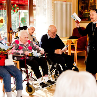 Eine Gruppe älterer Menschen sitzt in einem Raum. Eine Nonne steht vorne und spricht enthusiastisch, während einige Zuhörer ihre Blätter in den Händen halten. Die Atmosphäre ist lebhaft und gemeinschaftlich, mit einem Fokus auf Interaktion und Freude.