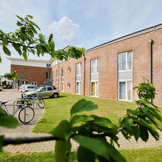 Das Bild zeigt ein mehrstöckiges Backsteingebäude mit zahlreichen Fenstern und einem Parkplatz davor. Im Vordergrund befinden sich Fahrräder und grüne Pflanzen. Der Himmel ist klar mit einigen Wolken.