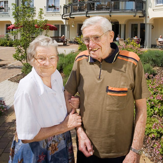 Ein älteres Paar steht lächelnd im Freien. Die Frau trägt ein weißes Oberteil und einen bunten Rock, während der Mann ein braunes T-Shirt mit Streifen hat. Sie halten sich an den Händen und wirken glücklich in ihrer Umgebung. Samen von Blumen und grünen Pflanzen sind zu sehen.