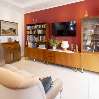 Ein gemütliches Wohnzimmer mit einem beigen Sofa und einer roten Wand. Auf der linken Seite steht ein Holzmöbelstück, daneben ein Bücherregal mit zahlreichen Büchern. An der Wand hängt ein Gemälde, und gegenüber steht ein Fernseher sowie ein Glaskasten mit Geschirr. Pflanzen und dekorative Objekte sind ebenfalls sichtbar.