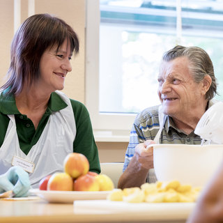 Eine Pflegerin und ein älterer Mann sitzen an einem Tisch und lächeln einander an. Vor ihnen stehen Äpfel und Schüsseln mit Zutaten. Die Atmosphäre ist freundlich und einladend, was auf eine gemeinschaftliche Aktivität hinweist.
