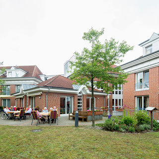 Eine Gruppe von älteren Menschen sitzt an Tischen im Freien vor einem Wohnheim. Einige Personen sind in Gespräche vertieft, während andere die Umgebung genießen. Umgeben von Bäumen und Gartenflächen vermittelt die Szene eine einladende, gesellige Atmosphäre.