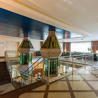 Eine helle und einladende Hotellobby mit einer Treppe und zwei speziell gestalteten Säulen, die jeweils ein Aquarium enthalten. Die Wände sind mit Fenstern, die Tageslicht hereinlassen, geschmückt. Die Lounge-Bereiche sind komfortabel eingerichtet und laden zum Entspannen ein.
