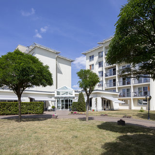 Das Bild zeigt ein modernes Gebäude mit mehreren Etagen und einem Eingangsbereich, umgeben von gepflegten Grünflächen und Bäumen. Der Himmel ist blau mit einigen Wolken, was eine freundliche und einladende Atmosphäre schafft.