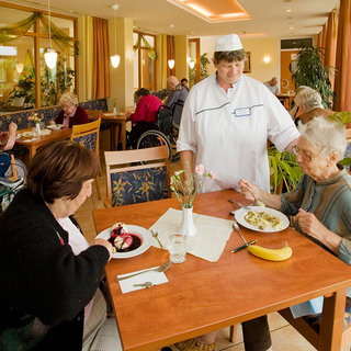 Eine Kellnerin mit einer weißen Uniform und einem Schirmmütze bedient zwei ältere Frauen an einem Tisch in einem Speisesaal. Eine Frau isst Nudeln, die andere hat ein Dessert vor sich. Im Hintergrund sind weitere Senioren zusammen. Raum wirkt hell und einladend.