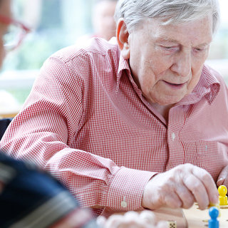 Ein älterer Mann mit grauen Haaren spielt konzentriert ein Brettspiel. Er sitzt an einem Tisch und bewegt eine Spielfigur. Im Hintergrund ist eine weitere Person, die nicht im Fokus steht. Die Atmosphäre wirkt freundlich und entspannt.